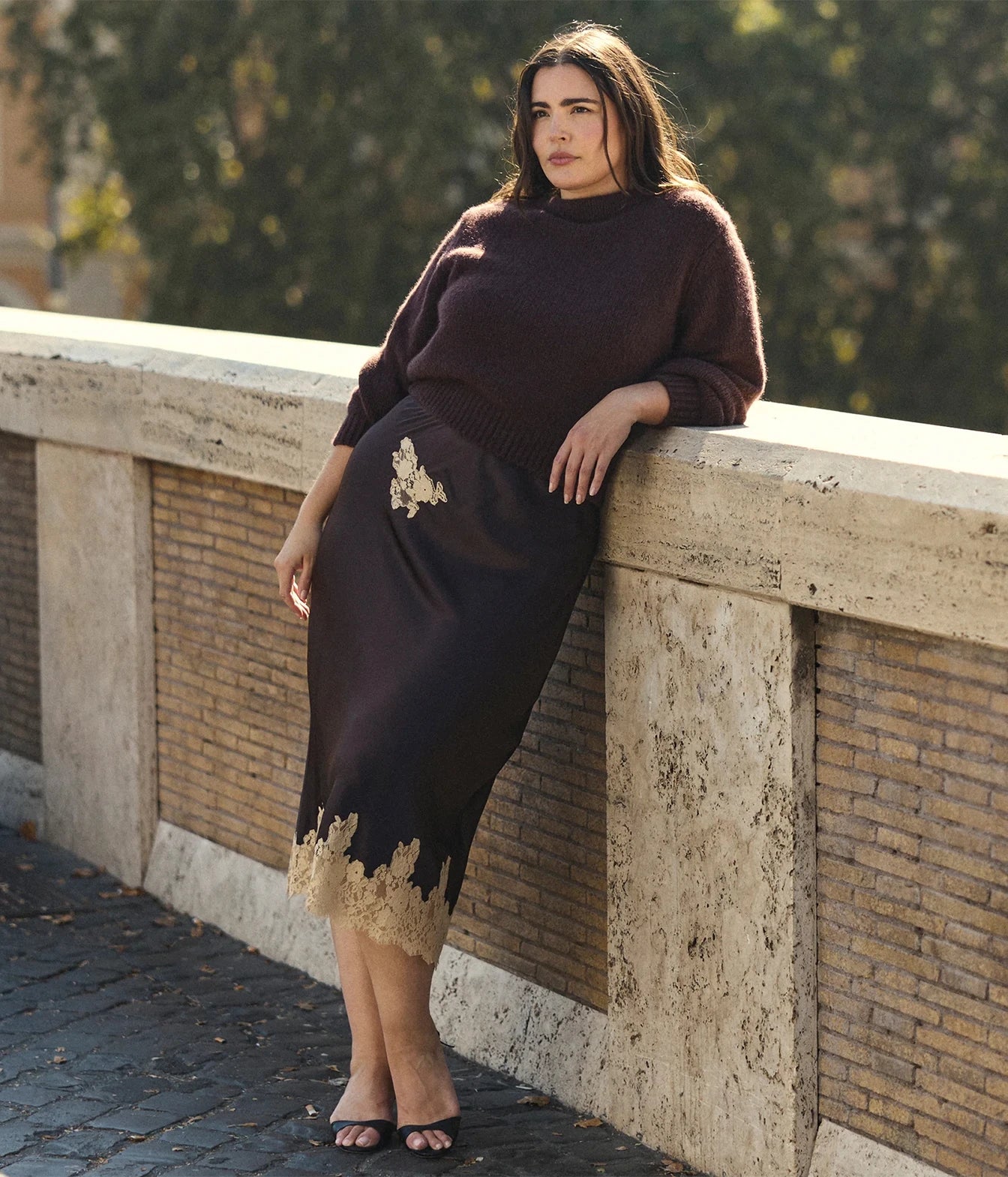 A woman leans against a stone bridge railing, wearing a dark brown mock-neck knit sweater and a matching dark brown midi skirt with gold lace trim and embroidery.