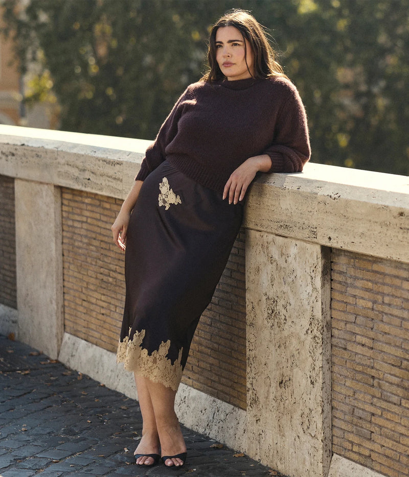 A woman leans against a stone bridge railing, wearing a dark brown mock-neck knit sweater and a matching dark brown midi skirt with gold lace trim and embroidery.