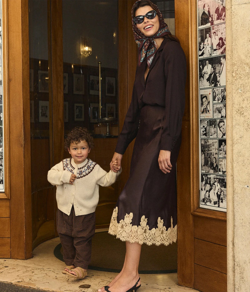 A woman in a dark brown long-sleeve dress with a beige lace hem and a patterned headscarf holds hands with a young boy wearing a cream sweater and brown pants, standing in a doorway.