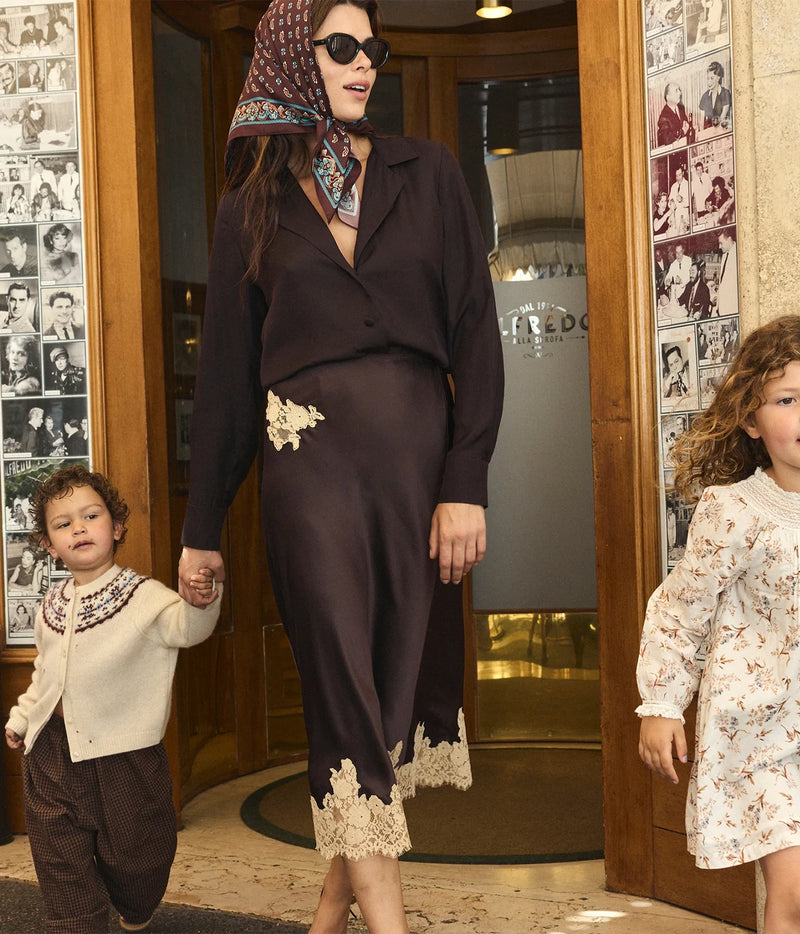 A woman wearing a dark brown long-sleeve button-up shirt and matching skirt with lace trim walks through a doorway with two children. The setting appears to be a boutique or interior hallway with framed photos on the walls.