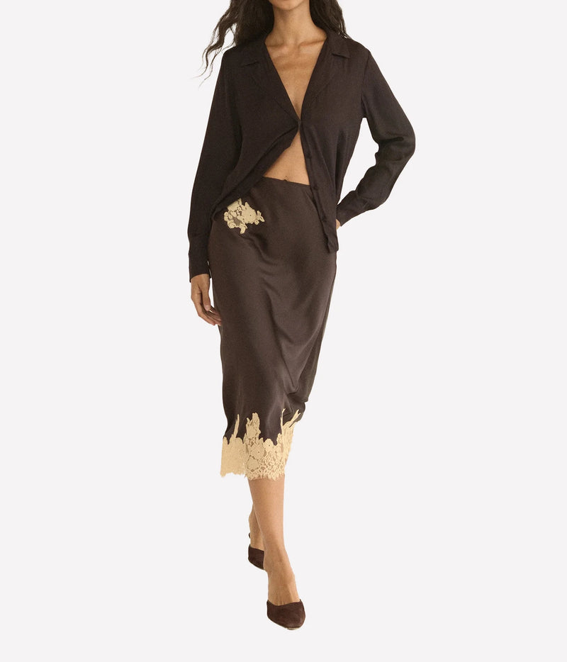 A model wearing a dark brown two-piece set consisting of a long-sleeve open-front jacket and a matching satin midi skirt with a beige floral applique and lace hem, set against a plain light gray background.