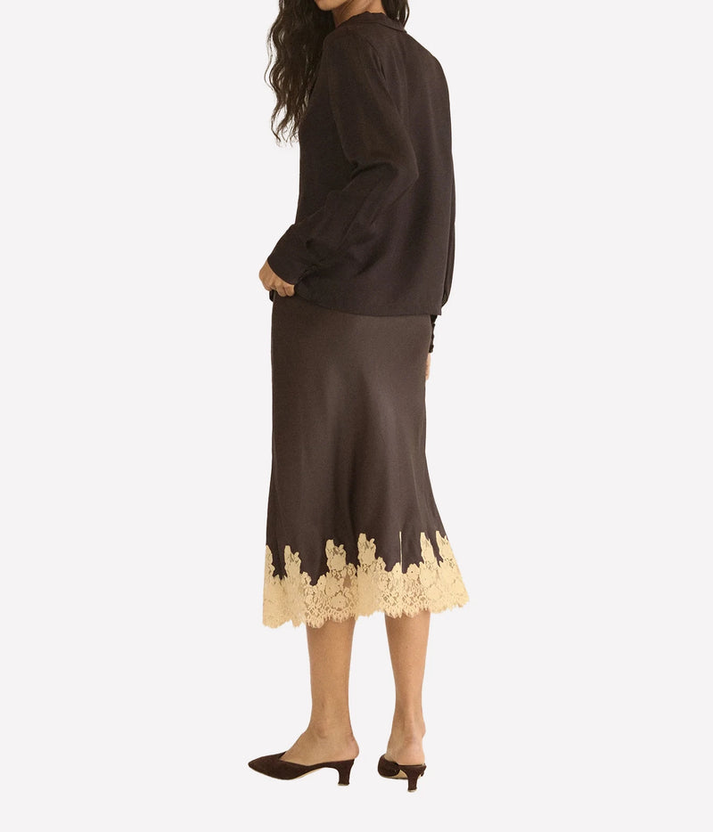 A woman shown from the back wearing a dark brown long-sleeve blouse and matching midi skirt with a beige floral lace hem, paired with dark brown mule heels, against a light gray studio background.
