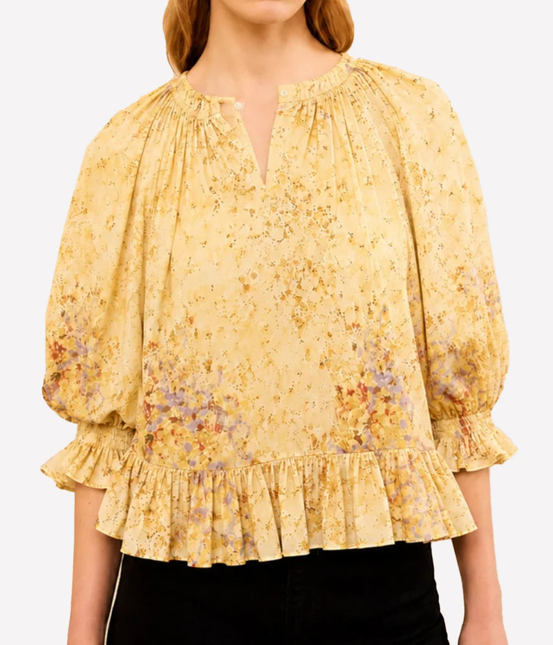 A woman wearing a yellow floral blouse with puff sleeves, a split neckline, and a ruffled hem, paired with black pants against a light gray studio background.