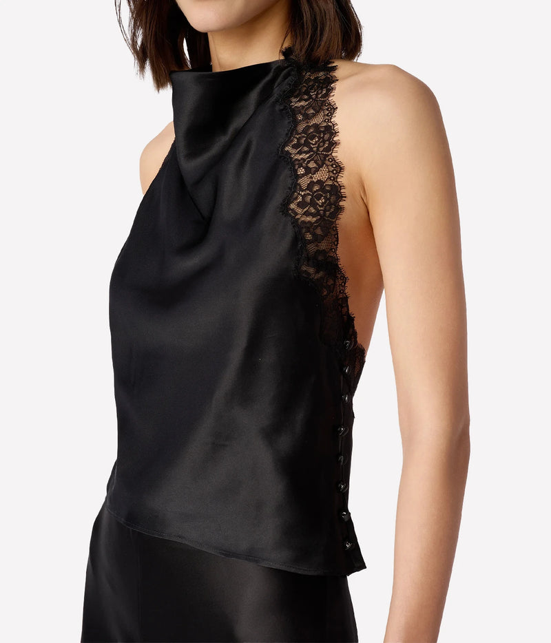 A close-up of a model wearing a black satin halter top with a high neck and a sheer black lace panel on the side, paired with a matching black satin skirt against a light gray background.
