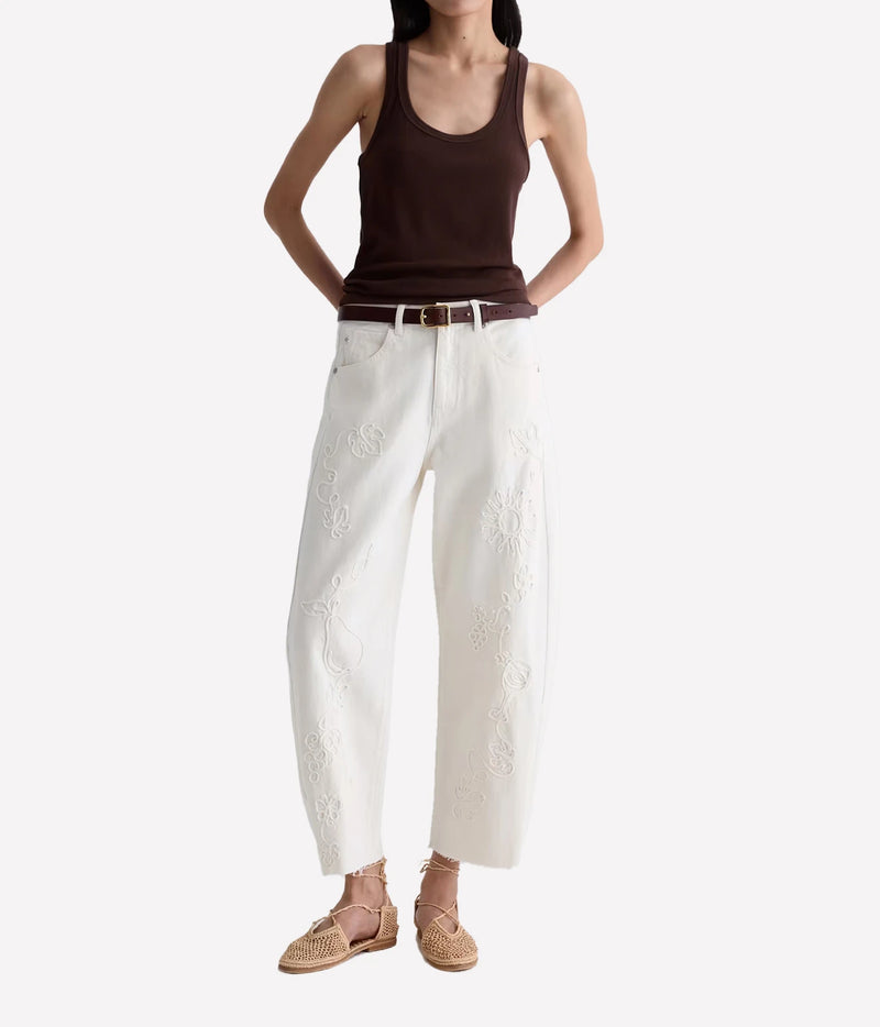A model is shown from the neck down wearing white wide-leg cropped pants with floral embroidery, styled with a dark brown ribbed tank top, a thin brown belt, and woven straw sandals against a plain white background.