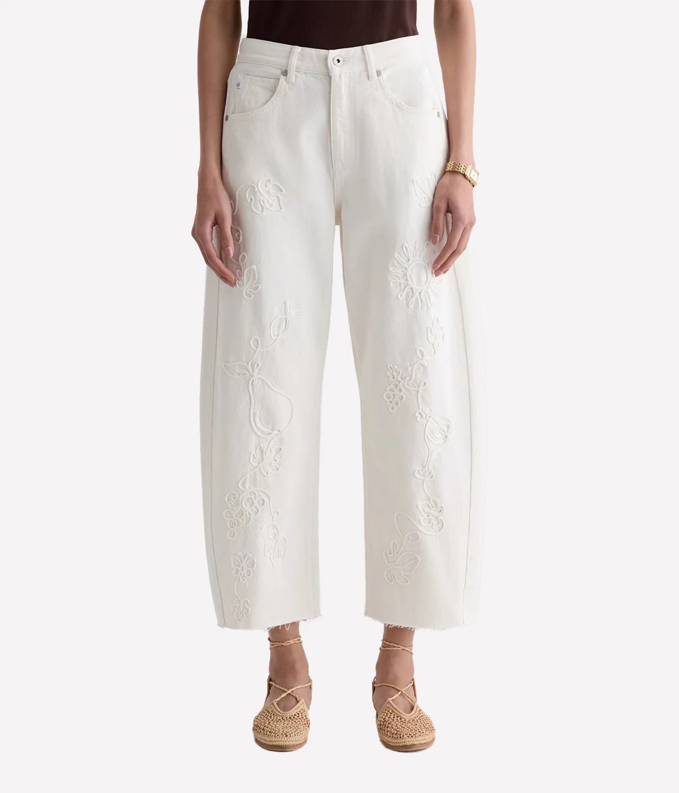 A model is wearing a pair of off-white cropped wide-leg jeans with intricate floral embroidery down the front of each leg, styled with woven espadrille sandals against a light gray background.