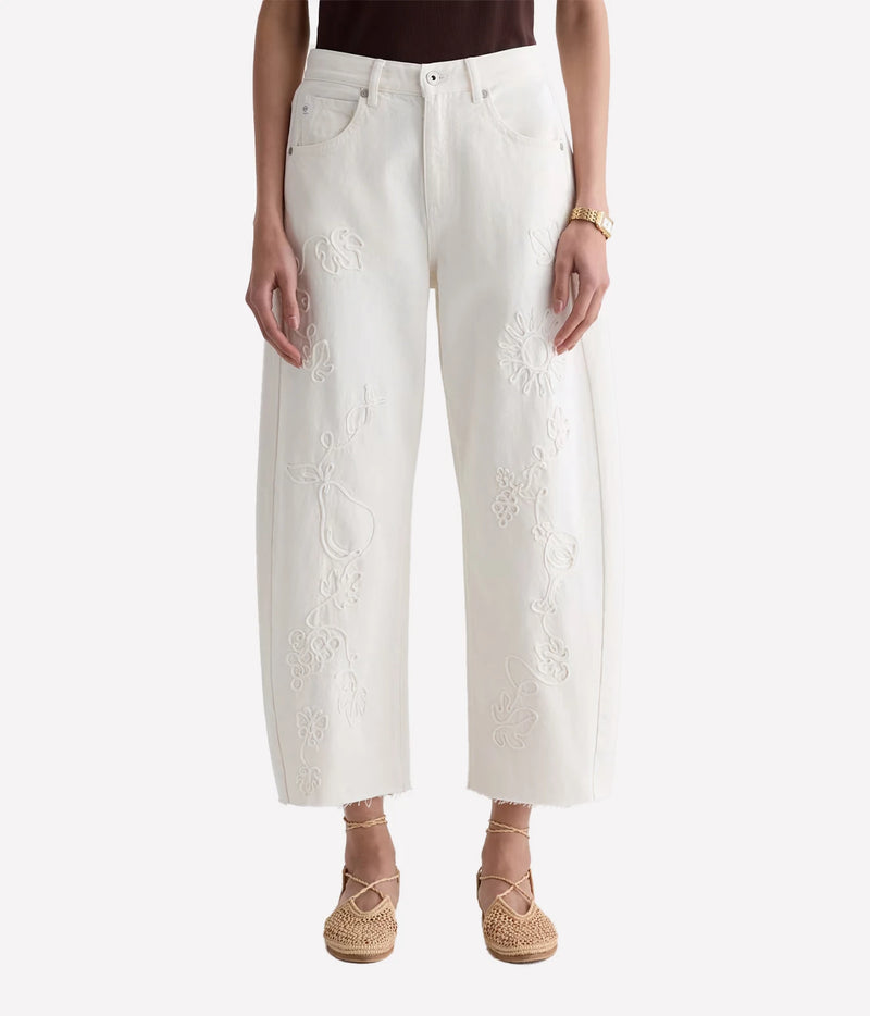 A model is wearing a pair of off-white cropped wide-leg jeans with intricate floral embroidery down the front of each leg, styled with woven espadrille sandals against a light gray background.