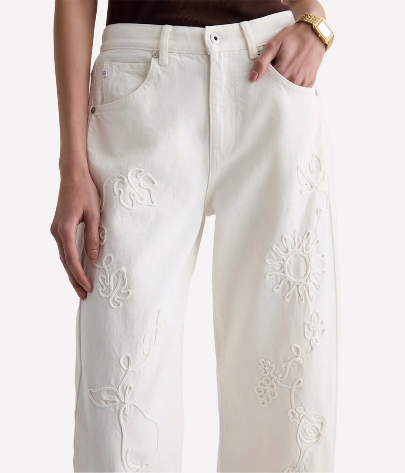 A close-up of a person wearing off-white embroidered jeans with floral cutwork details, shown from the waist down against a plain light background.