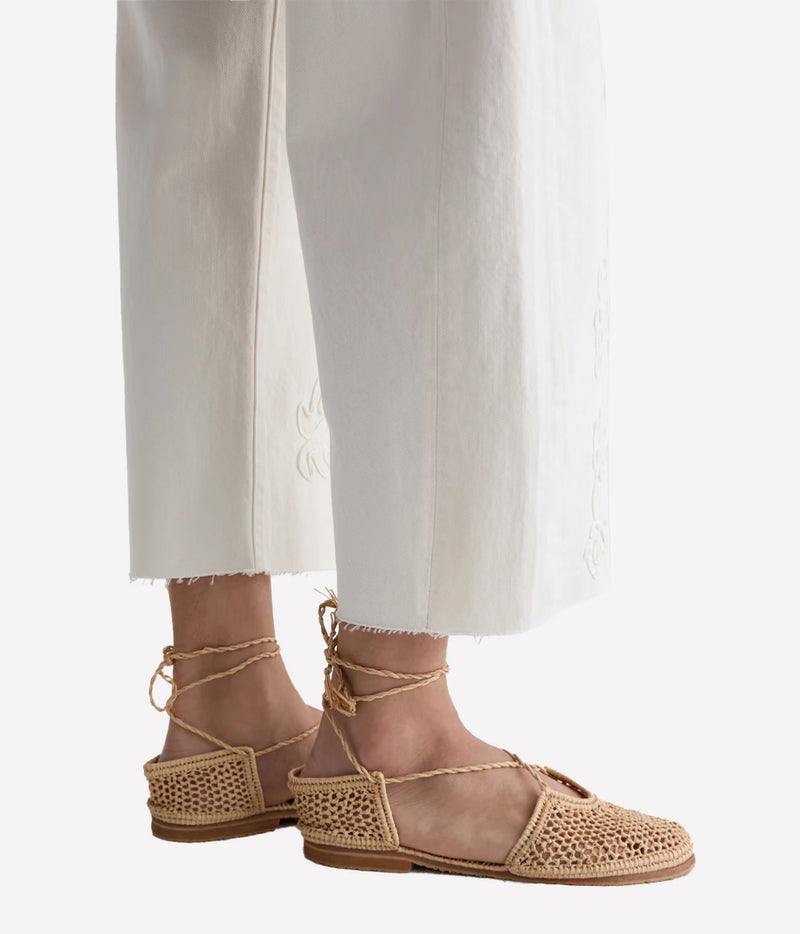 A pair of natural woven espadrille sandals with lace-up ankle straps, worn with white cropped trousers against a light gray studio background.