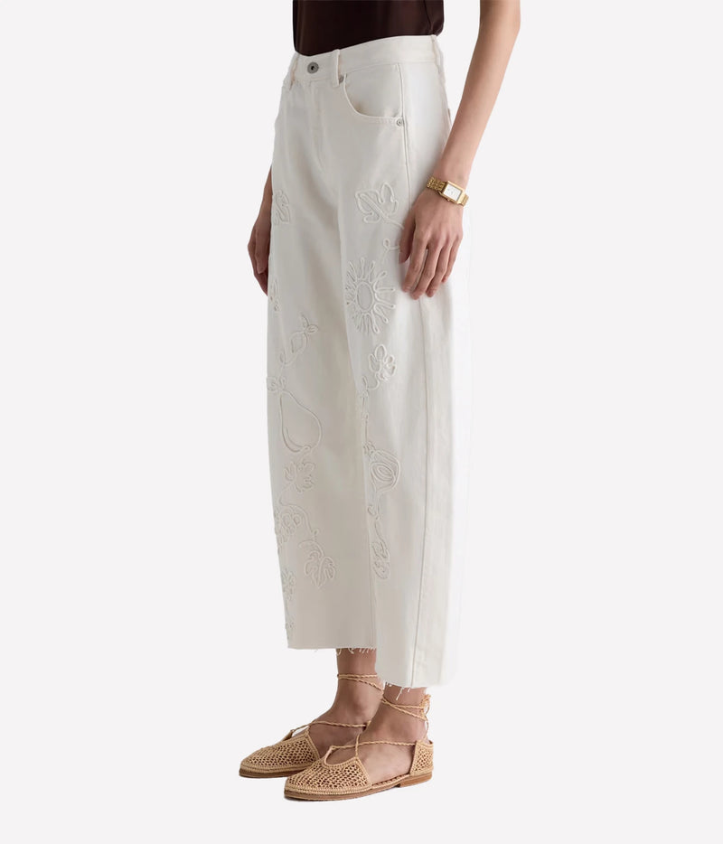 A model wearing off-white embroidered wide-leg cropped pants with floral and paisley stitching, paired with beige woven flat sandals and a gold watch, against a light gray studio background.