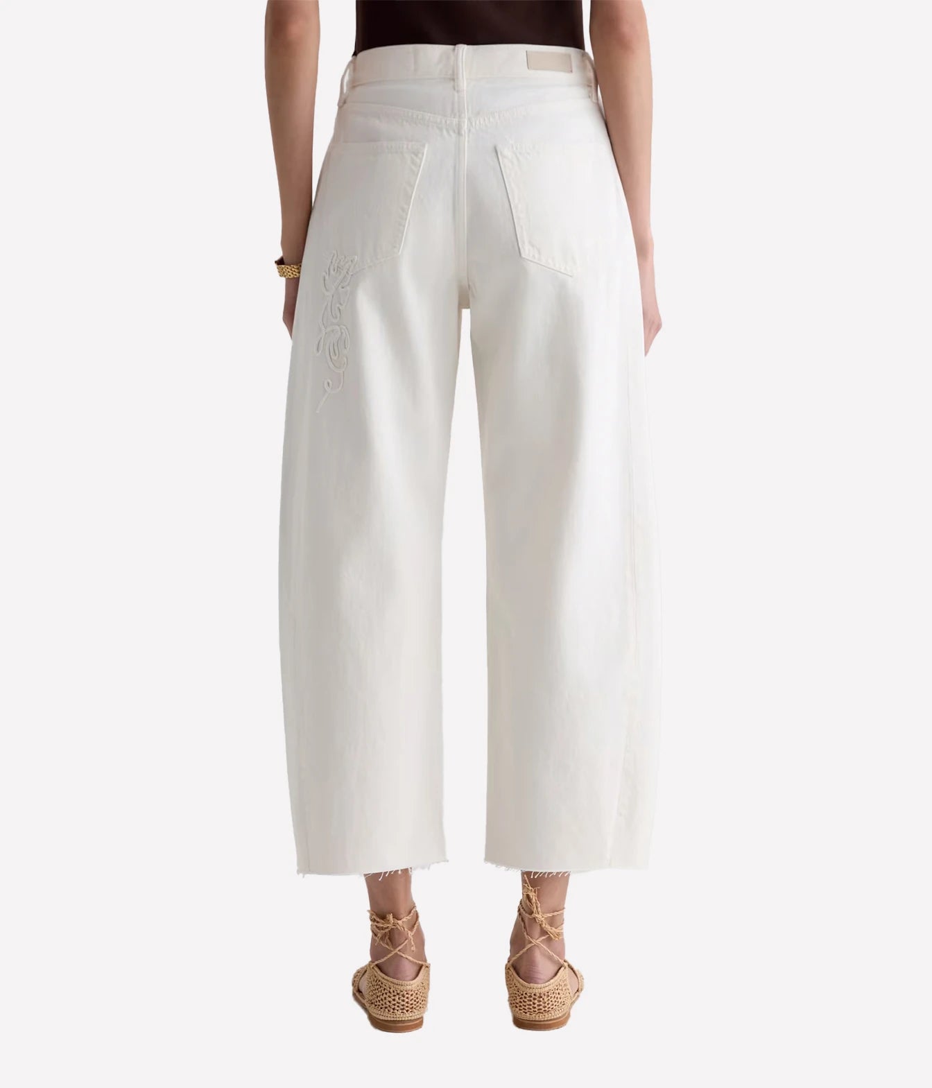 A model shown from the back wearing off-white cropped wide-leg jeans with embroidered detailing on the left leg, paired with a dark top and beige lace-up espadrille sandals against a light gray background.