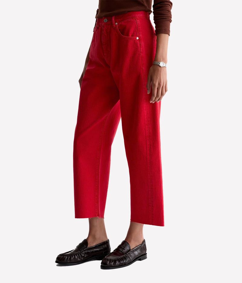 Three-quarter front view of a model wearing the AG Hattie Crop in Recording Red with a brown knit top, silver watch and dark brown loafers, showing the curved leg shape