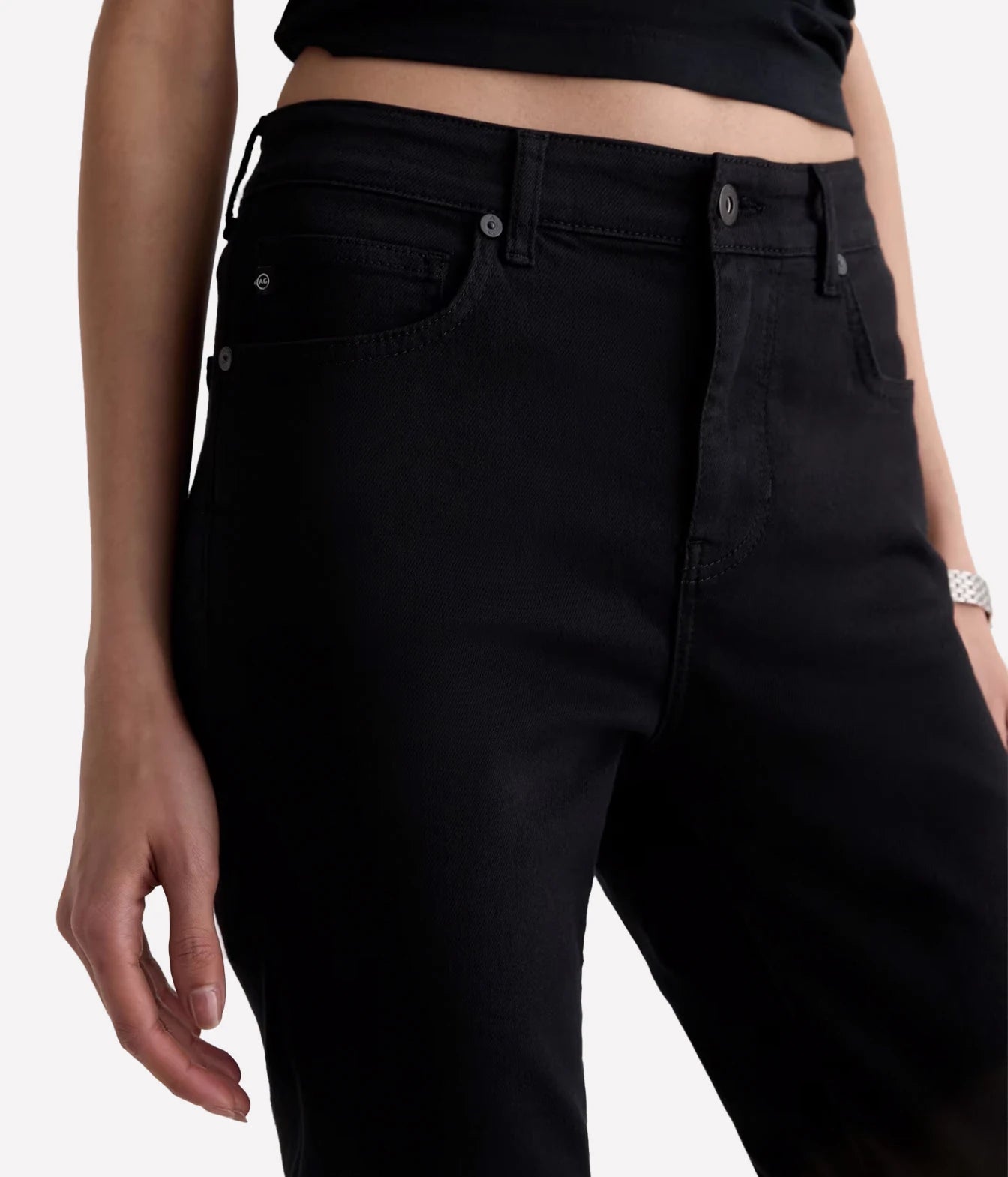 Close-up of a model wearing black denim jeans with a cropped black top, showing the front waistband, belt loops, and front pocket details against a light gray background.
