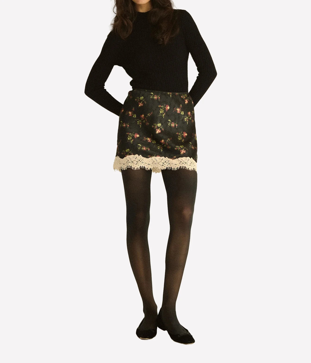 Full-length front view of a model wearing the DÔEN Giulietta Skirt In Noir Hillside Thistles with a black knit top, sheer black tights and black flats