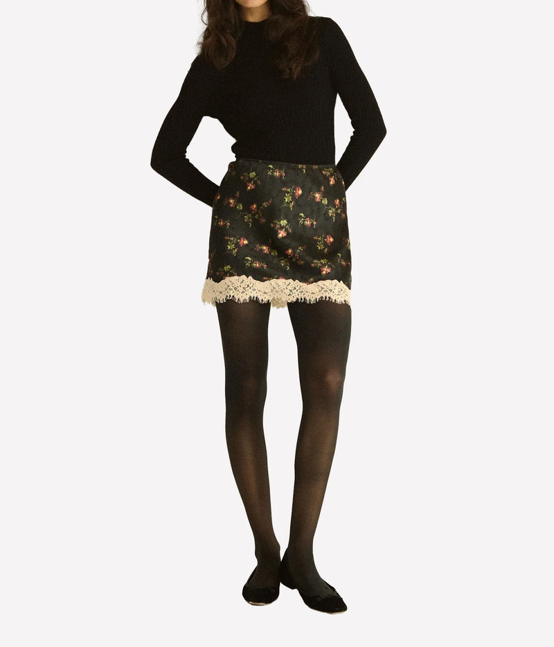 Full-length front view of a model wearing the DÔEN Giulietta Skirt In Noir Hillside Thistles with a black knit top, sheer black tights and black flats