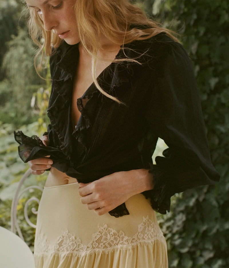 A woman outdoors wearing a black ruffled blouse and a cream lace-trimmed skirt, adjusting the blouse with one hand.