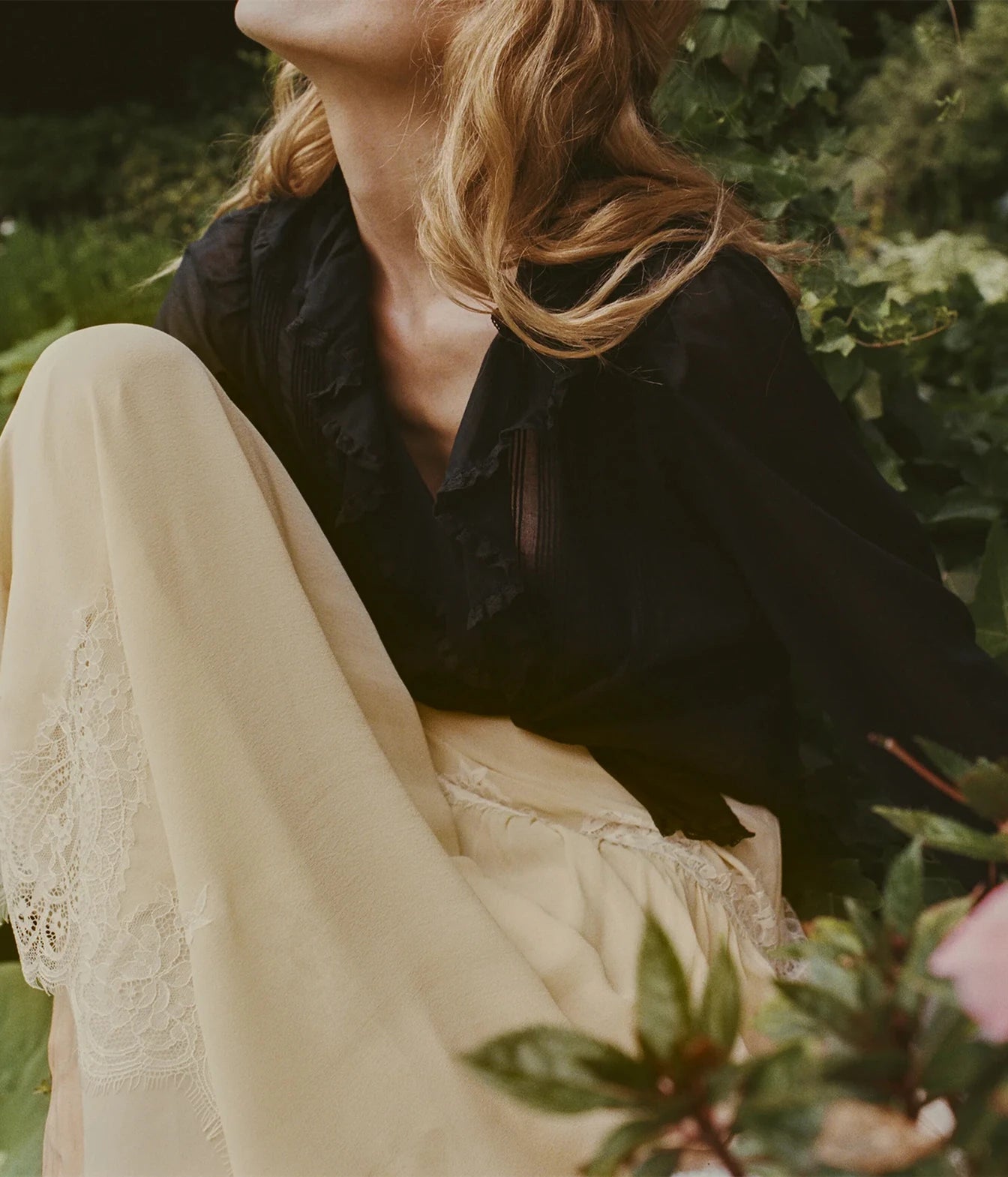 A woman outdoors wearing a black ruffled blouse and cream wide-leg trousers with lace trim, seated among green plants.