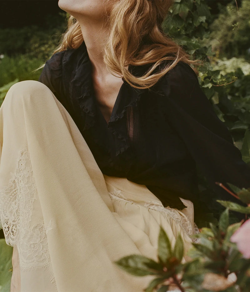 A woman outdoors wearing a black ruffled blouse and cream wide-leg trousers with lace trim, seated among green plants.