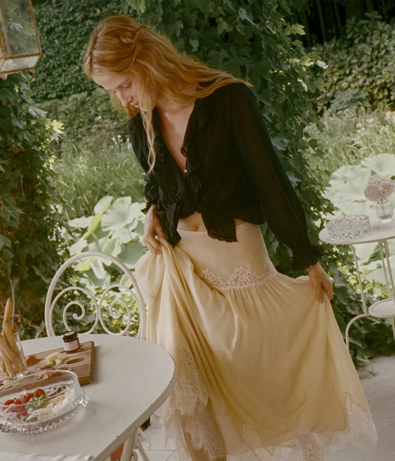 A woman stands on an outdoor patio surrounded by green foliage, wearing a sheer black long-sleeve top and a flowing cream-colored skirt with lace trim. She is holding out the skirt to show its drape.