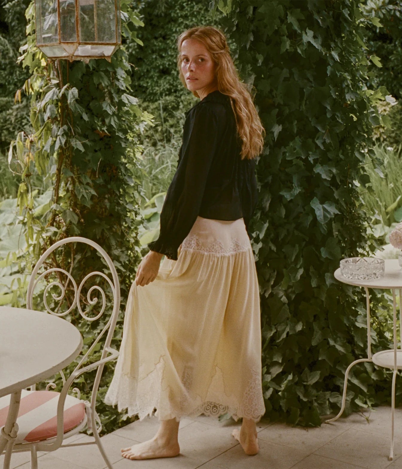 A woman stands barefoot on a patio surrounded by green ivy, wearing a black long-sleeve blouse and a cream-colored embroidered skirt.