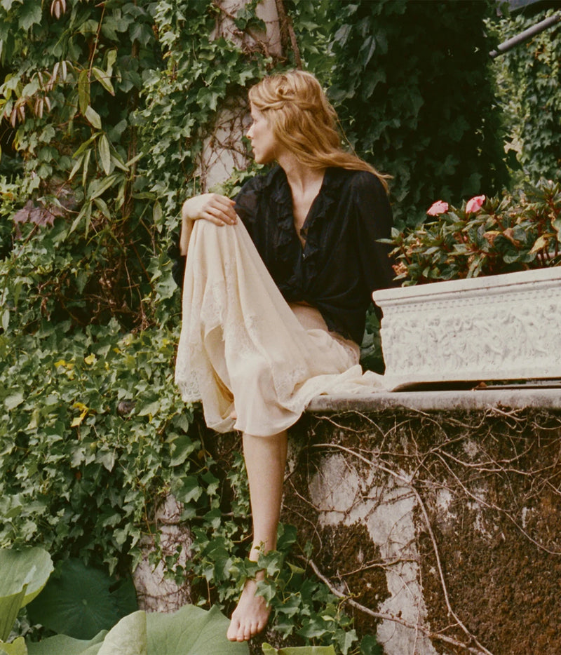 A woman with blonde hair sits barefoot on a stone ledge surrounded by lush green ivy and a planter box, wearing a black blouse and a flowing cream skirt.