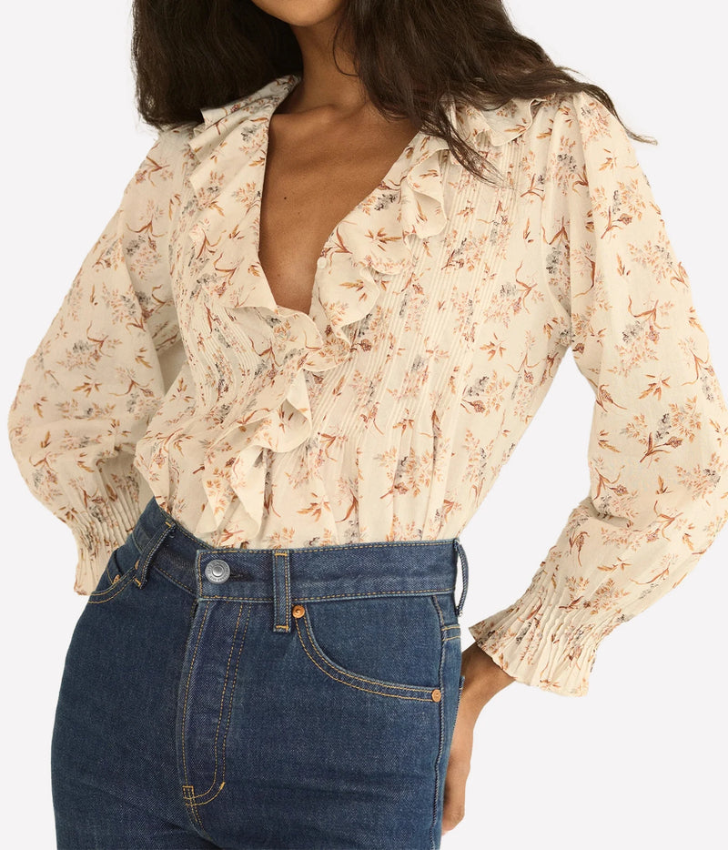 A woman wearing a beige long-sleeve blouse with a delicate brown and orange floral print, ruffled trim, and smocked cuffs, paired with dark blue jeans against a light gray background.