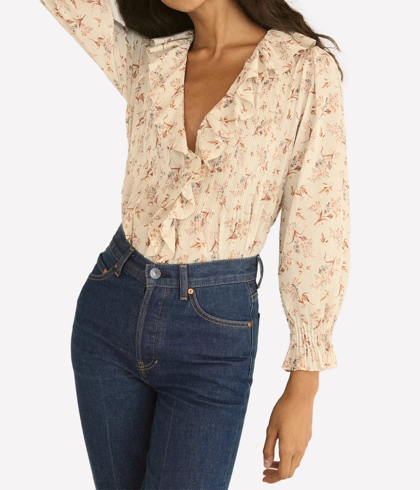 A woman wearing a cream-colored long-sleeve blouse with a delicate pink and brown floral print, paired with dark blue jeans, shown against a plain light gray background.