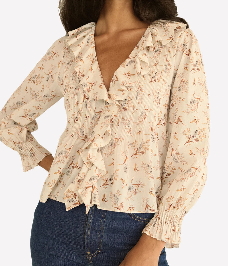 A woman wearing a cream-colored long-sleeve blouse with a delicate orange and brown floral print, ruffle details, and smocked texture, paired with blue jeans against a light gray background.
