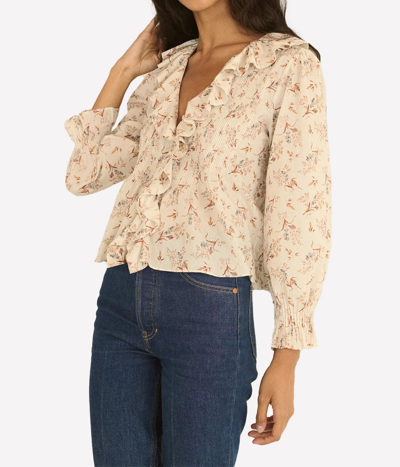 A woman wearing a beige long-sleeve blouse with a delicate pink and brown floral print, ruffled trim, and a split neckline, paired with dark blue jeans against a light gray background.