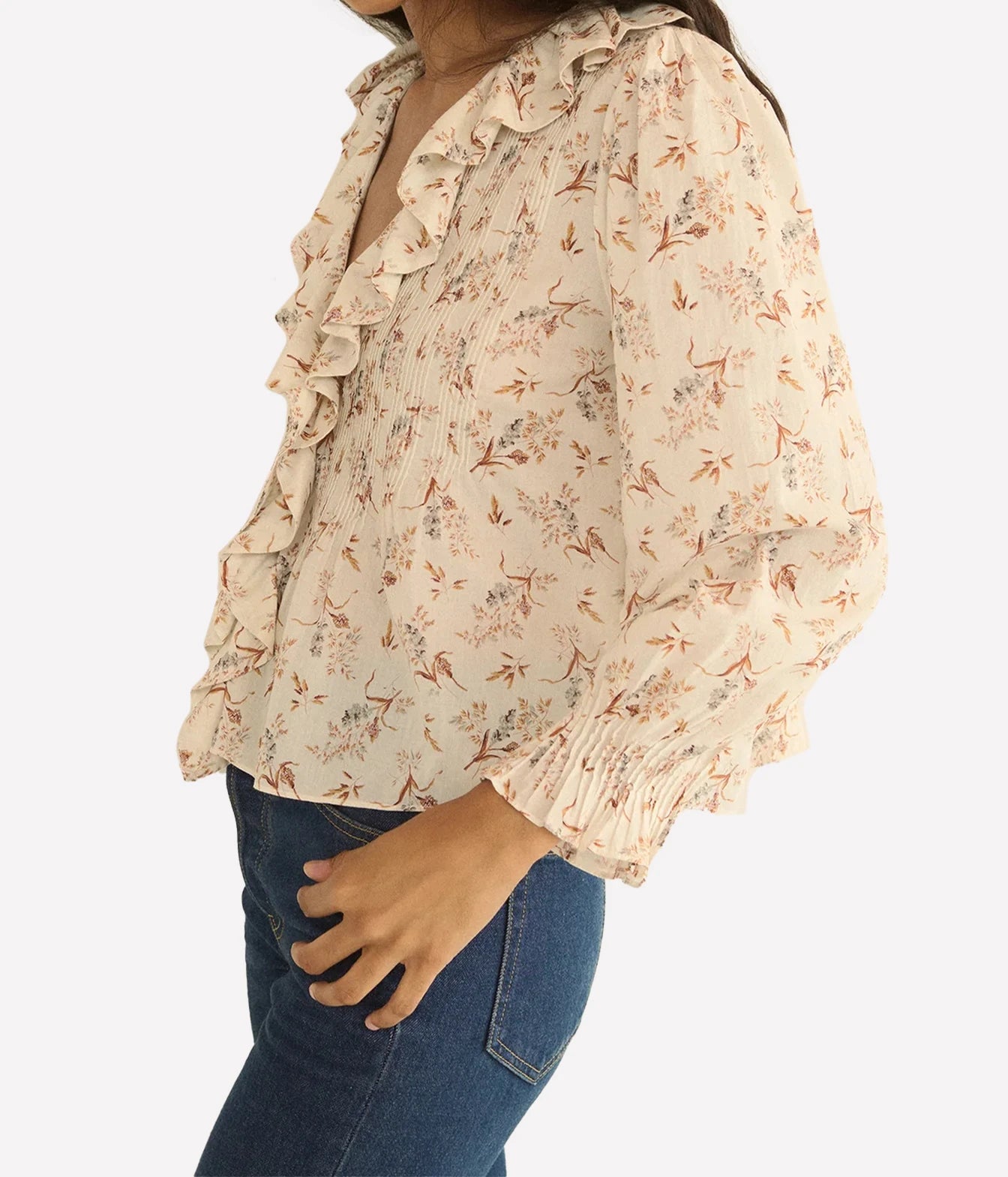 A woman wearing a cream-colored blouse with a delicate orange and brown floral print, ruffled trim along the collar and sleeve, and a smocked waist. She is paired with blue jeans against a clean white background.