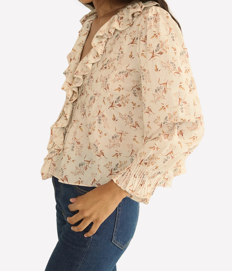 A woman wearing a cream-colored blouse with a delicate orange and brown floral print, ruffled trim along the collar and sleeve, and a smocked waist. She is paired with blue jeans against a clean white background.
