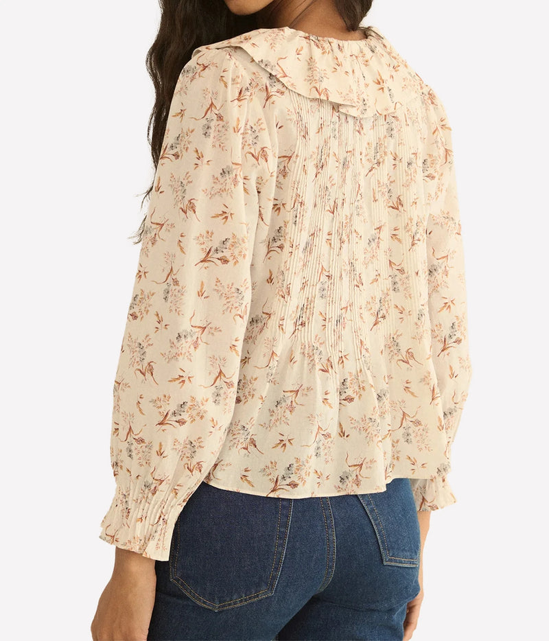 A woman wearing a cream-colored long-sleeve blouse with a delicate brown and orange floral pattern, paired with blue jeans, shown from a three-quarters back view against a light gray background.