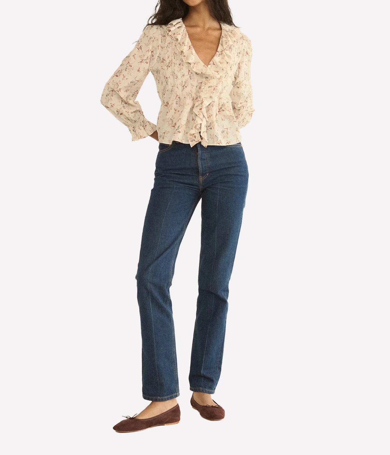 A model wearing a cream floral long-sleeve blouse with ruffles, blue straight-leg jeans, and brown flat shoes, photographed against a light gray studio background.