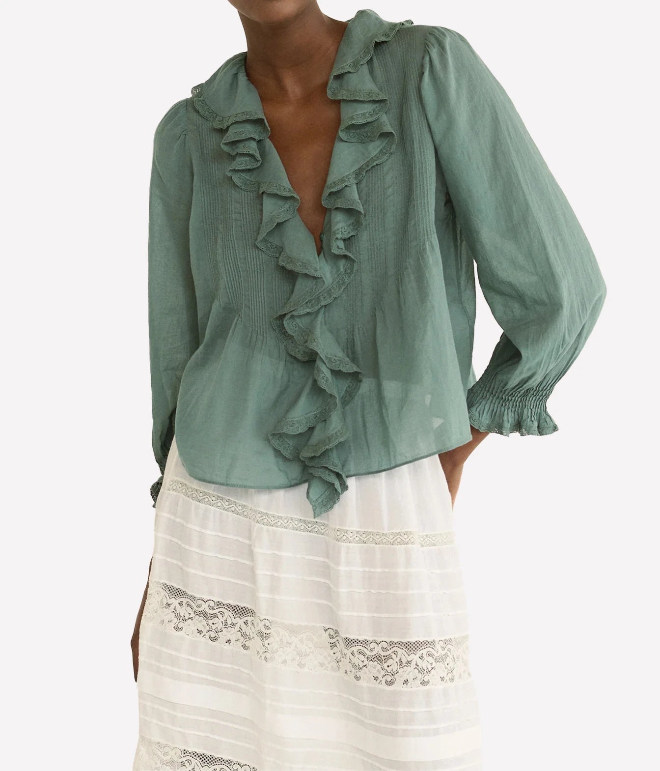 A woman wearing a sage green long-sleeve blouse with ruffle trim and a white skirt, shown against a plain light background.