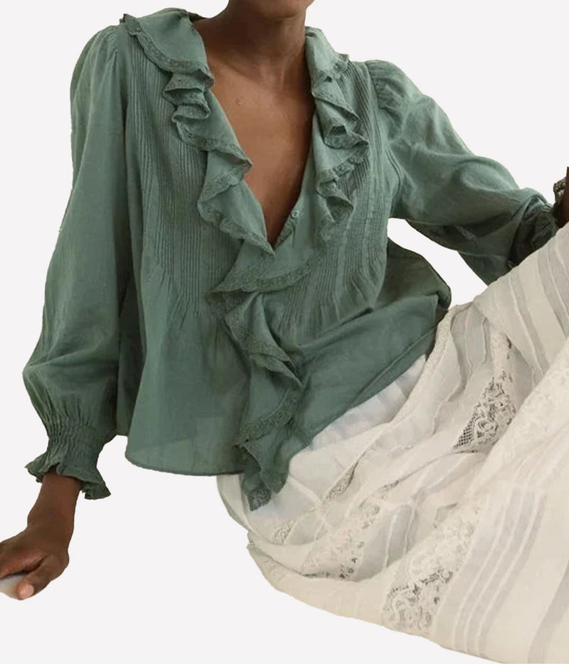 A woman wearing a sage green long-sleeve blouse with layered ruffles and a smocked neckline, paired with a white skirt, against a plain white background.
