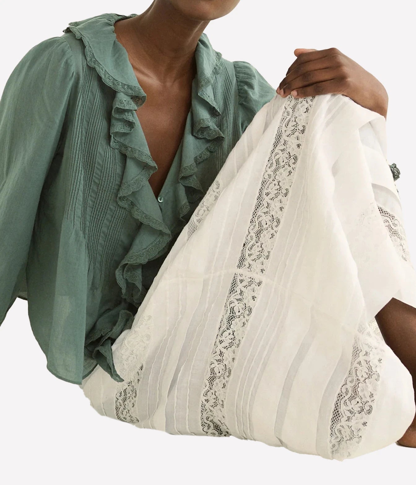 A close-up of a person wearing a sage green ruffled blouse and holding a white sheer fabric panel with black floral lace trim, against a plain light background.