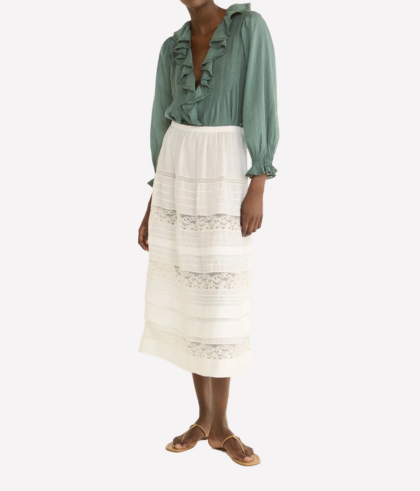 A model wearing a sage green long-sleeve blouse with ruffles and a high-waisted white midi skirt featuring horizontal lace trim, paired with tan flat sandals, against a light gray background.