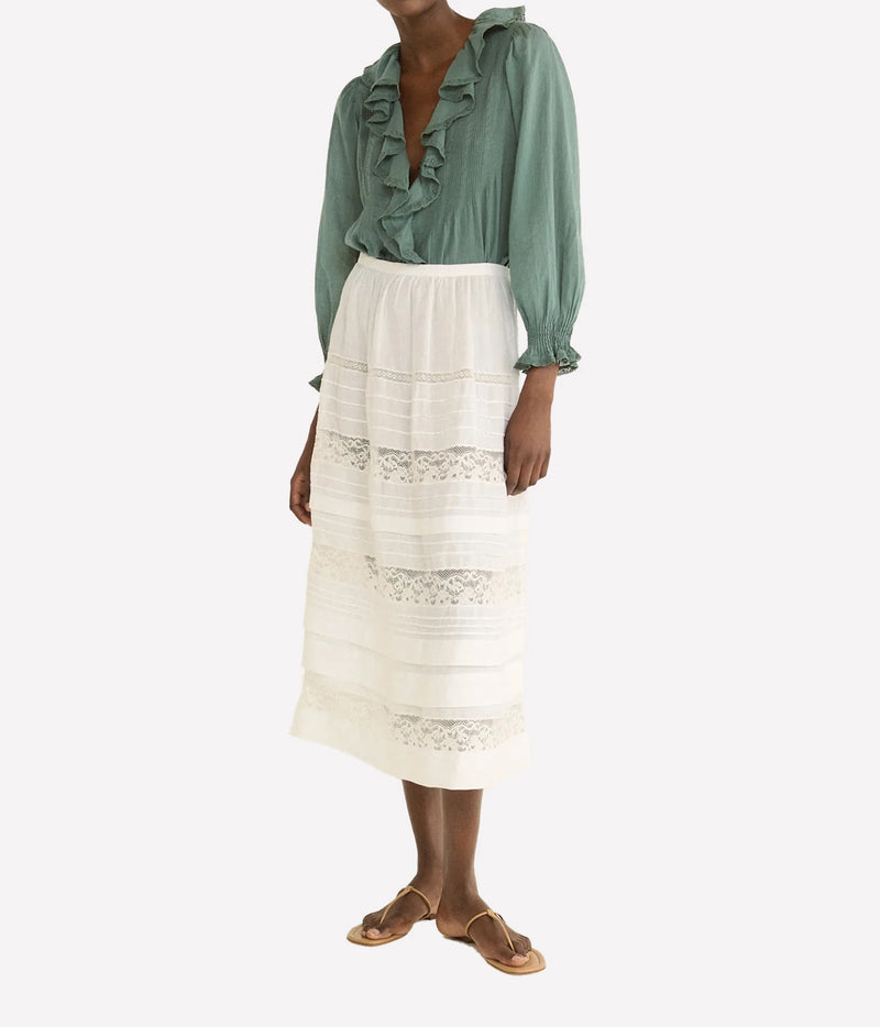 A model wearing a sage green long-sleeve blouse with ruffles and a high-waisted white midi skirt featuring horizontal lace trim, paired with tan flat sandals, against a light gray background.
