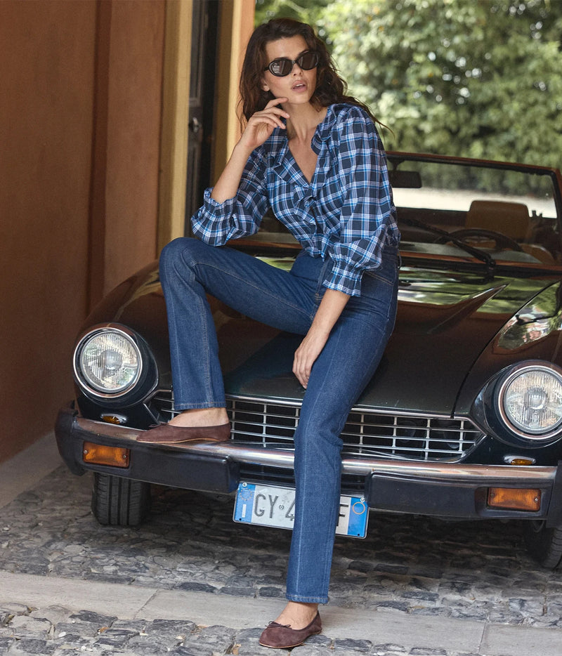 A woman wearing a blue and white plaid button-up shirt and dark blue jeans sits on the front bumper of a vintage black convertible car outdoors.