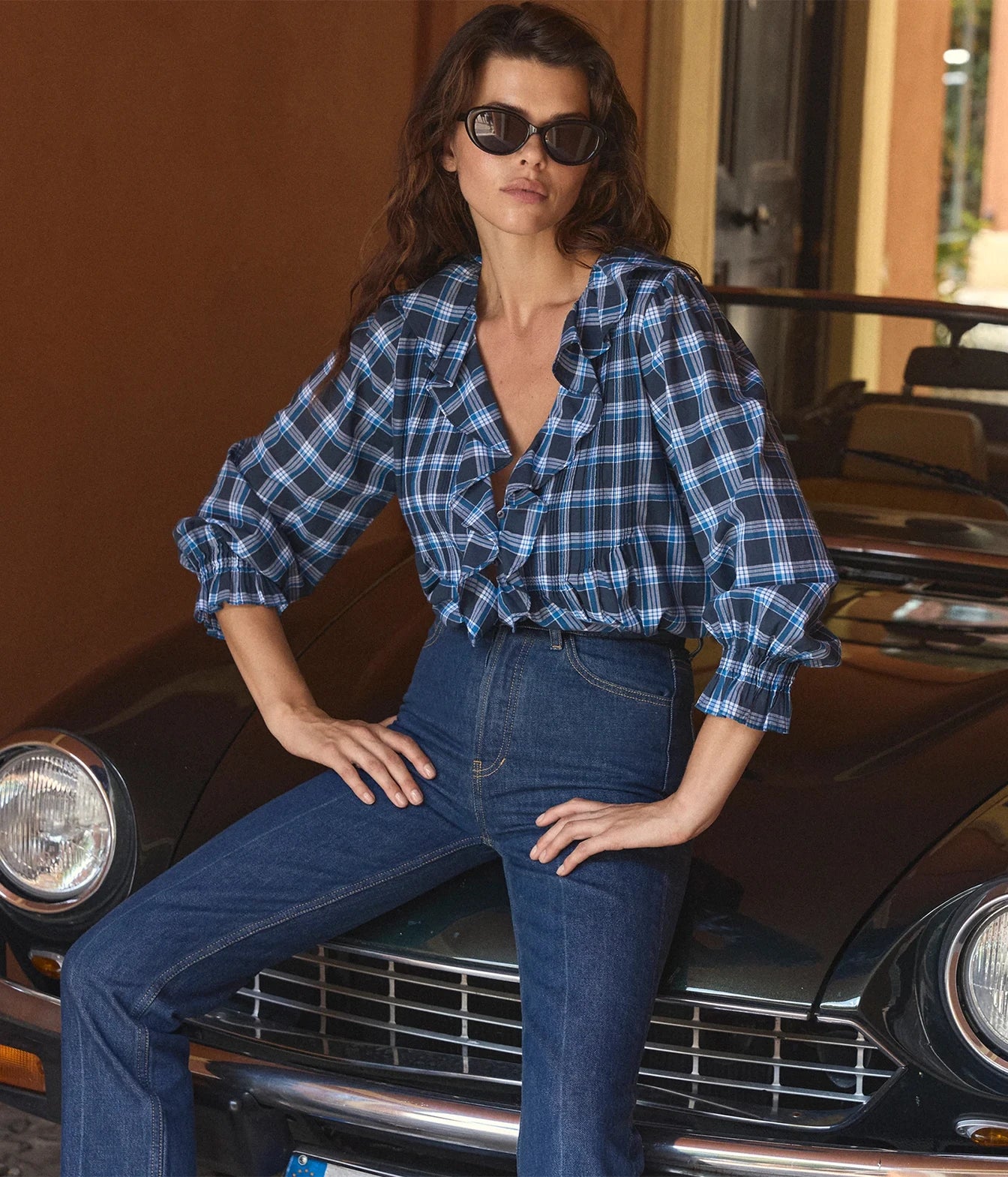 A woman wearing a blue and black plaid ruffled shirt and dark blue jeans sits on the front of a vintage black car in a warm-toned setting.