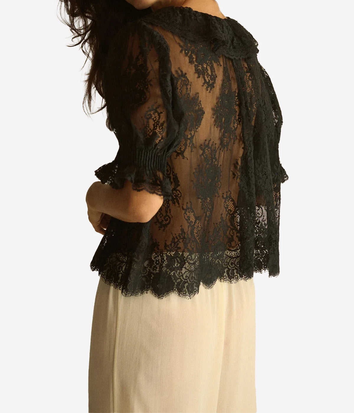 Back view of a model wearing the DÔEN Lace Henri Top In Black with cream trousers, showing the sheer lace back, ruffled sleeve cuffs and scalloped hem