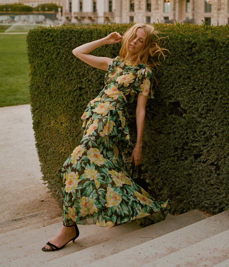 Full-length view of a model leaning against a hedge in the DÔEN Hesperia Dress In Hanbury Garden with black ankle-strap sandals, showing the tiered maxi skirt