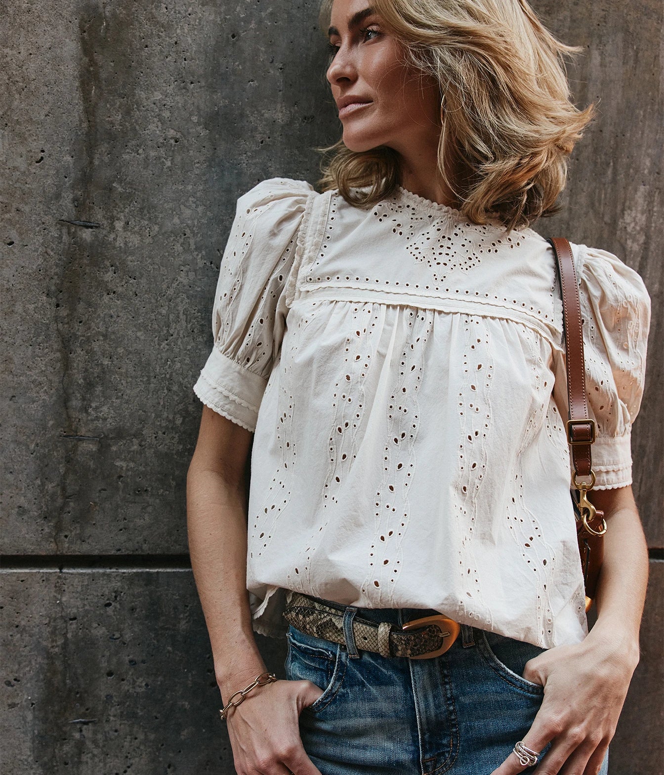 A woman wearing a cream eyelet short-sleeve blouse with puff sleeves, paired with blue jeans and a brown leather shoulder bag, standing against a textured gray concrete wall.