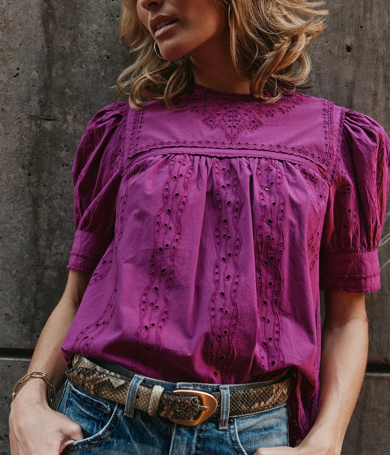 A woman wearing a magenta-purple eyelet blouse with puff sleeves and decorative embroidery, paired with blue jeans and a snakeskin belt, standing against a textured concrete wall.