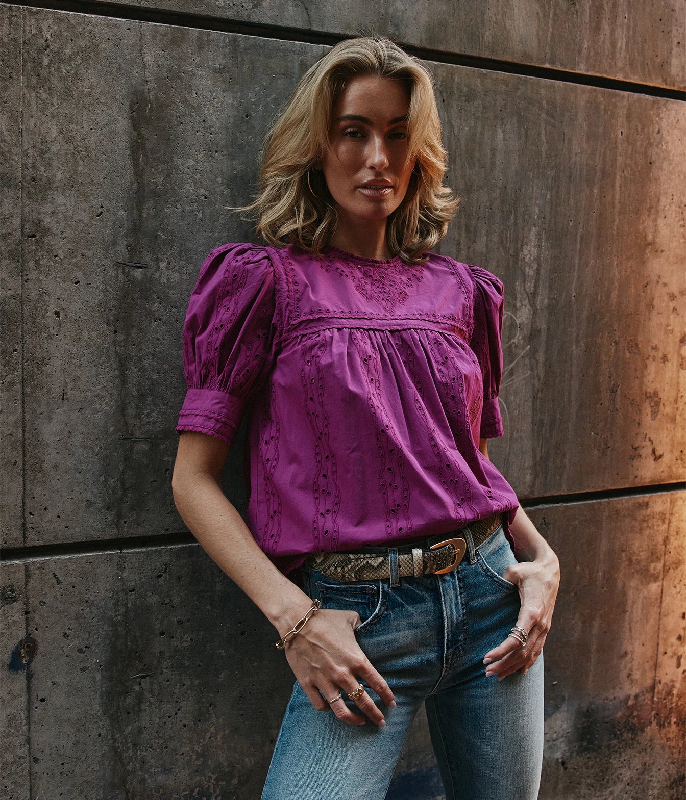 A woman wearing a purple short-sleeve eyelet blouse with puff sleeves, paired with blue jeans and a patterned belt, posing against a rustic concrete wall.