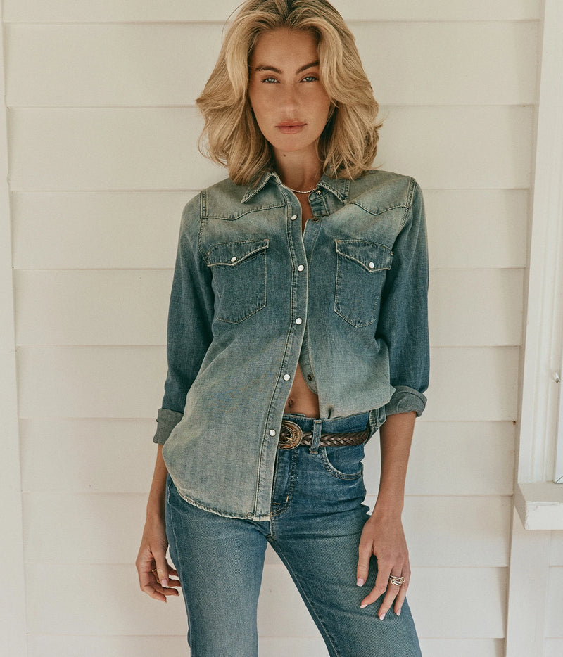 Nili Lotan Celia Jean In Classic Wash, worn on model with a denim western shirt tucked in and a brown belt, standing by a white weatherboard wall