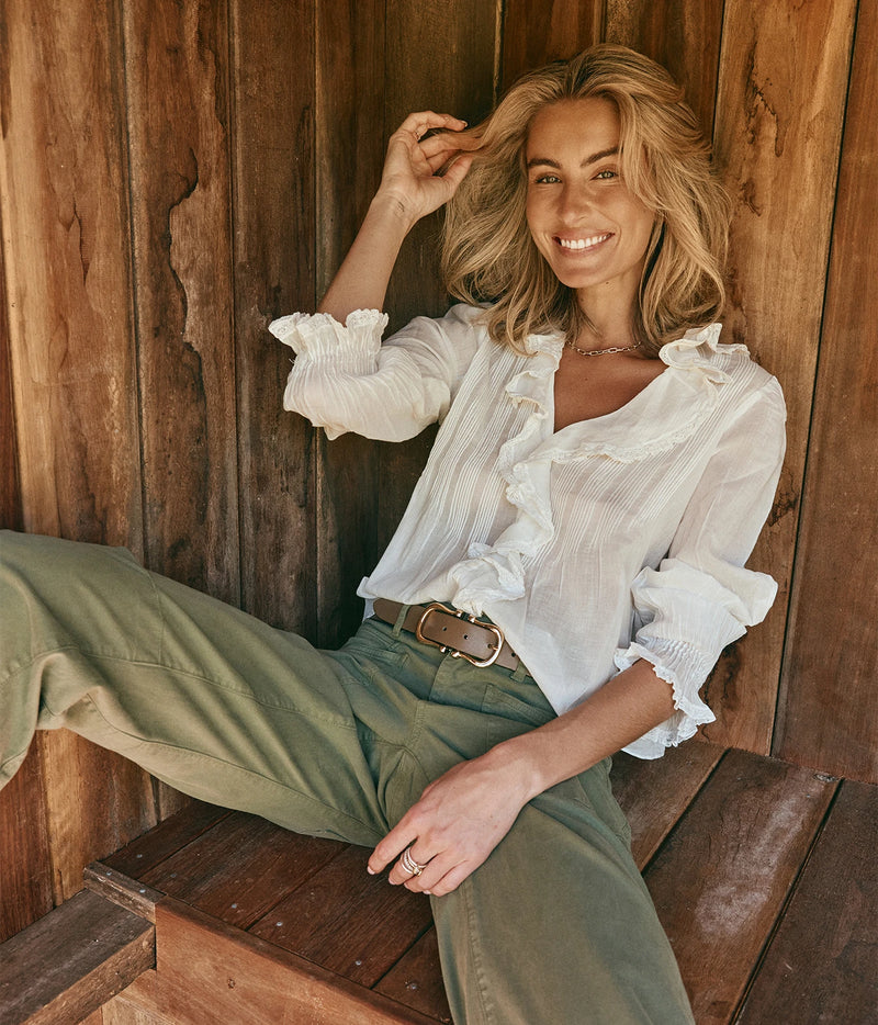 DÔEN Hardy Top In Salt, worn on model with olive trousers and a woven tan belt, seated on a wooden bench, showing the ruffled collar and cuffs