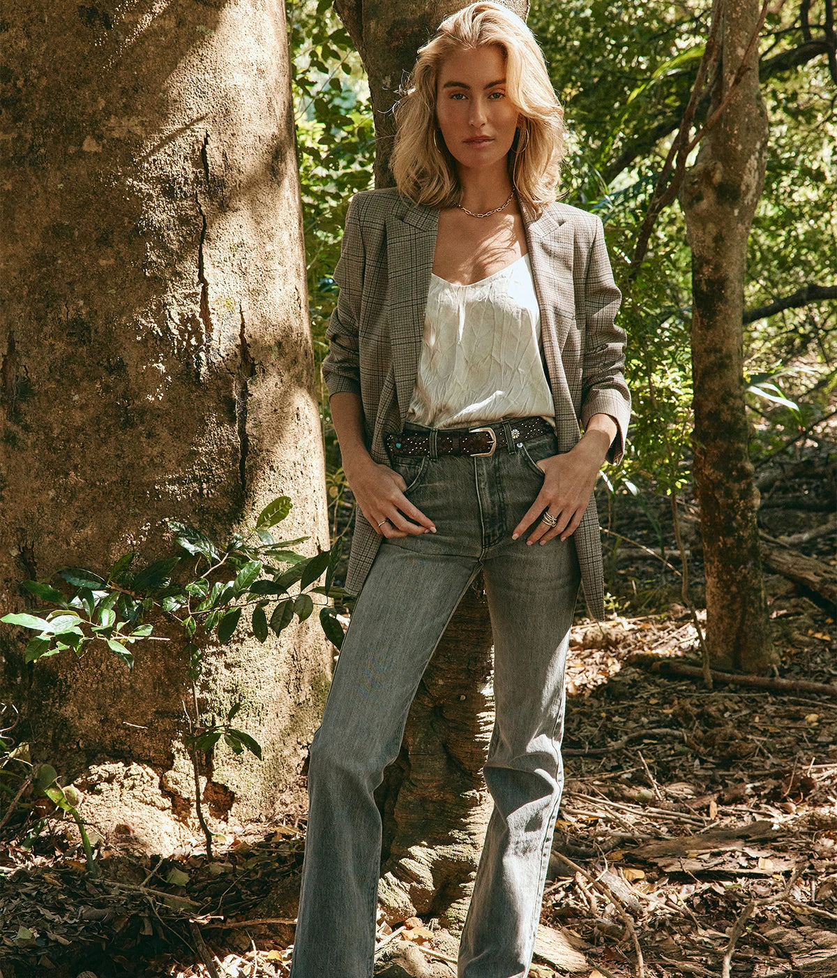 Calexico Fawcett Belt In T.Moro, worn on model through grey straight-leg jeans with a satin cami and check blazer, standing among trees