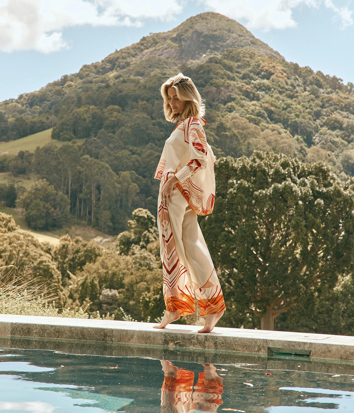 Raquel Diniz Bahia Shirt In Ivory Pink Tangerine Red, side view on model with the matching Sofia Pants, walking along the edge of a pool
