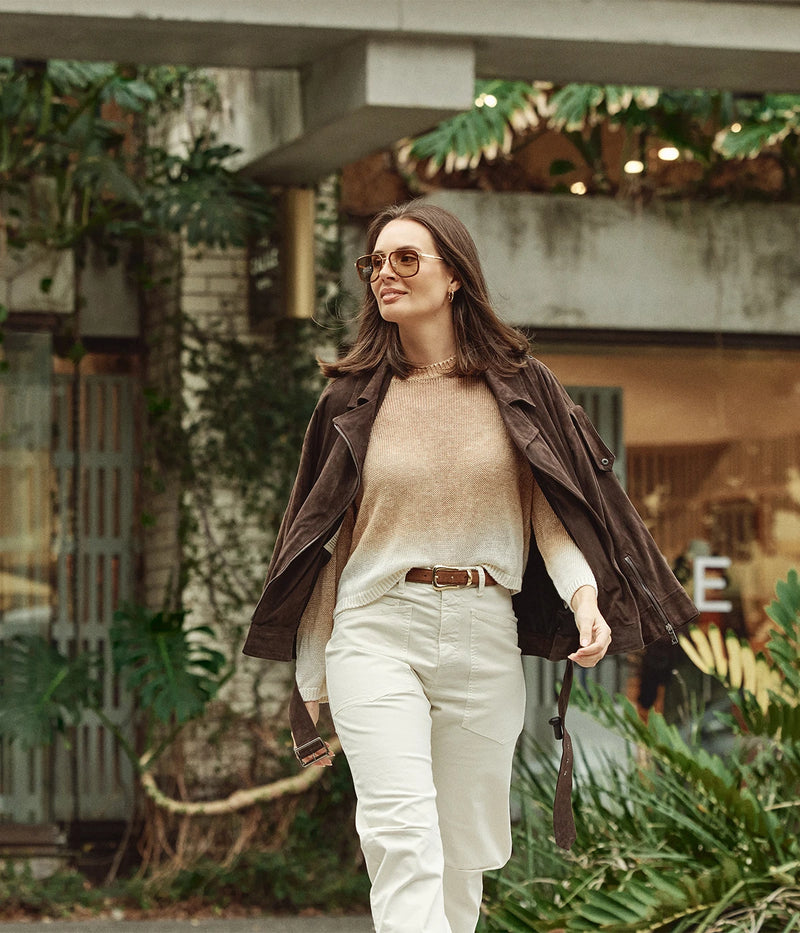 A woman walking outdoors wearing a dark brown leather jacket, a beige knit sweater, cream pants, and round sunglasses, set against a blurred urban background with greenery and building elements.